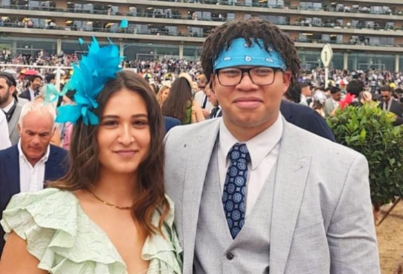 Alina wearing a blue fascinator and Dylan in a grey suit at Royal Ascot by the trackside rail