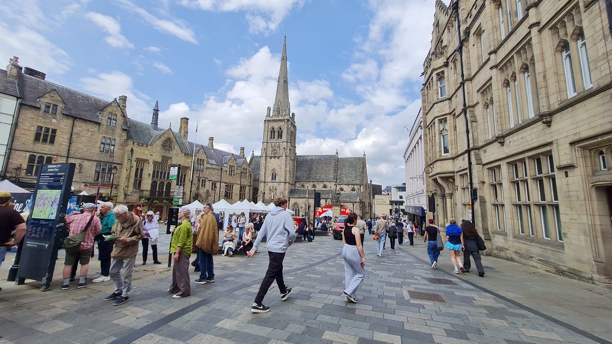 Durham Market Place busy with people and market stalls, St Nicholas’ Church behind
