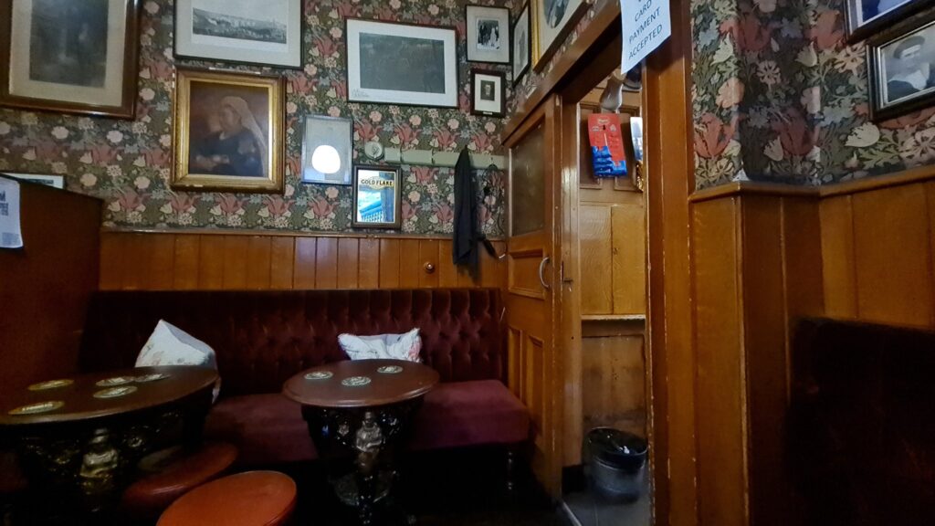 The snug at the Victoria Inn in Durham — a red button-back banquette, a cast-iron table and framed pictures hung on floral wallpaper