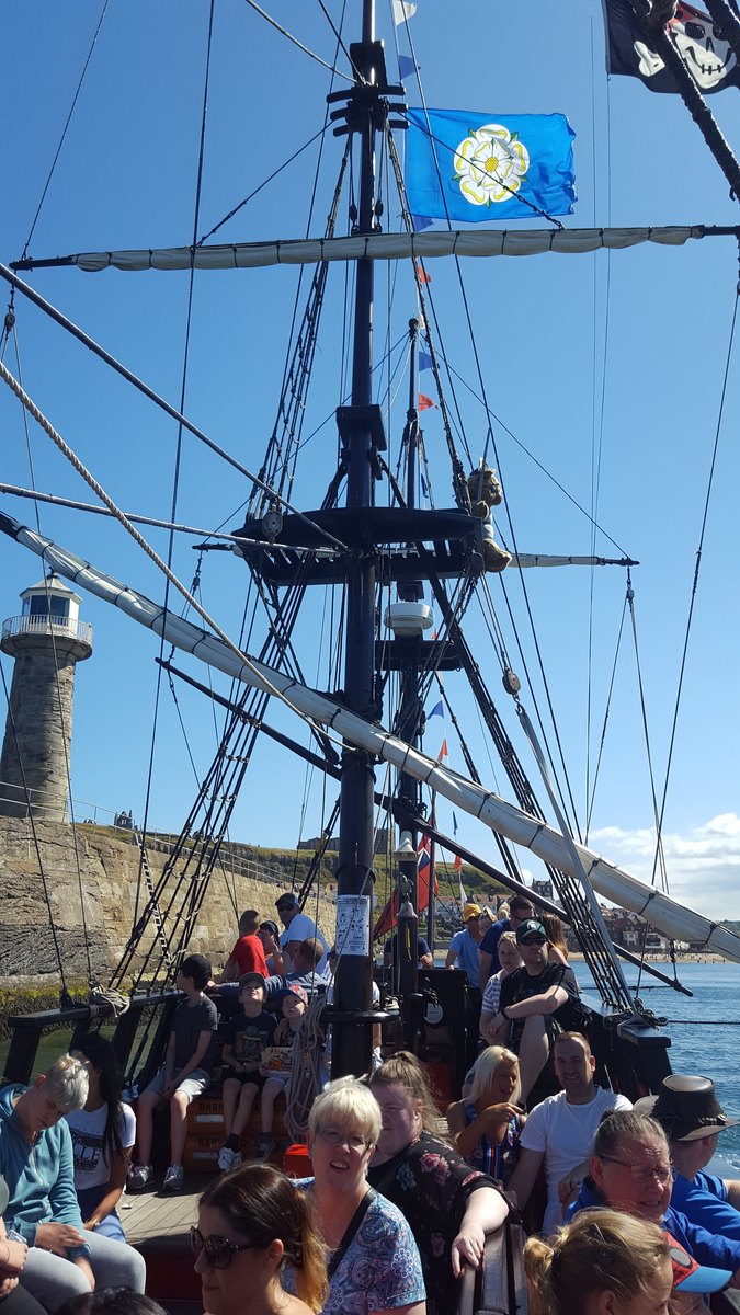 Looking up the masts and rigging of the Bark Endeavour from the deck in Whitby, with passengers sitting below and the pier lighthouse behind